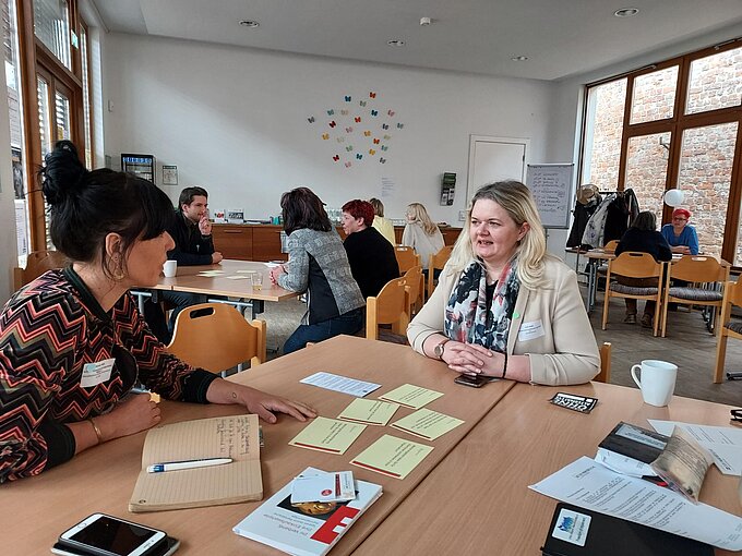 Zwei Frauen in einem großen Raum im Gespräch. Im Hintergrund weitere Gesprächssituationen.