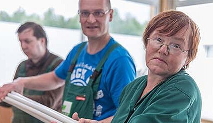 Drei Personen stehen in einem hellen Raum und halten gemeinsam lange Rohre. Sie tragen Arbeitskleidung und blicken direkt in die Kamera. Die Atmosphäre wirkt kollegial und engagiert.