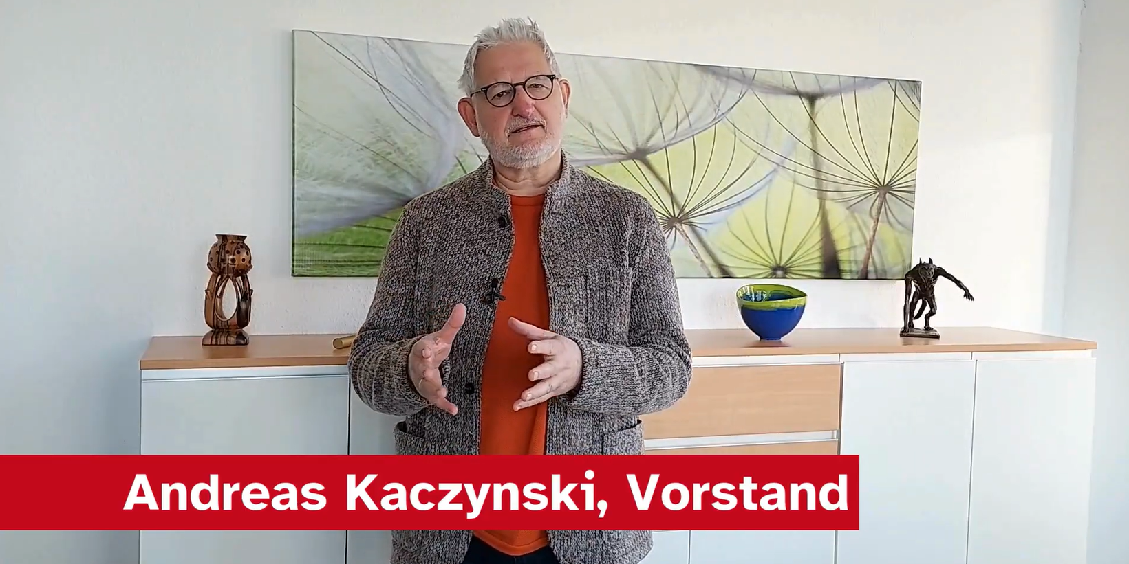 Ein Mann namens Andreas Kaczynski steht in einem hellen Raum vor einem Sideboard. Er trägt eine graue Jacke und ein oranges T-Shirt. Im Hintergrund ist ein großes Dekobild mit Pflanzenmotiven sowie einige dekorative Objekte auf dem Sideboard zu sehen.