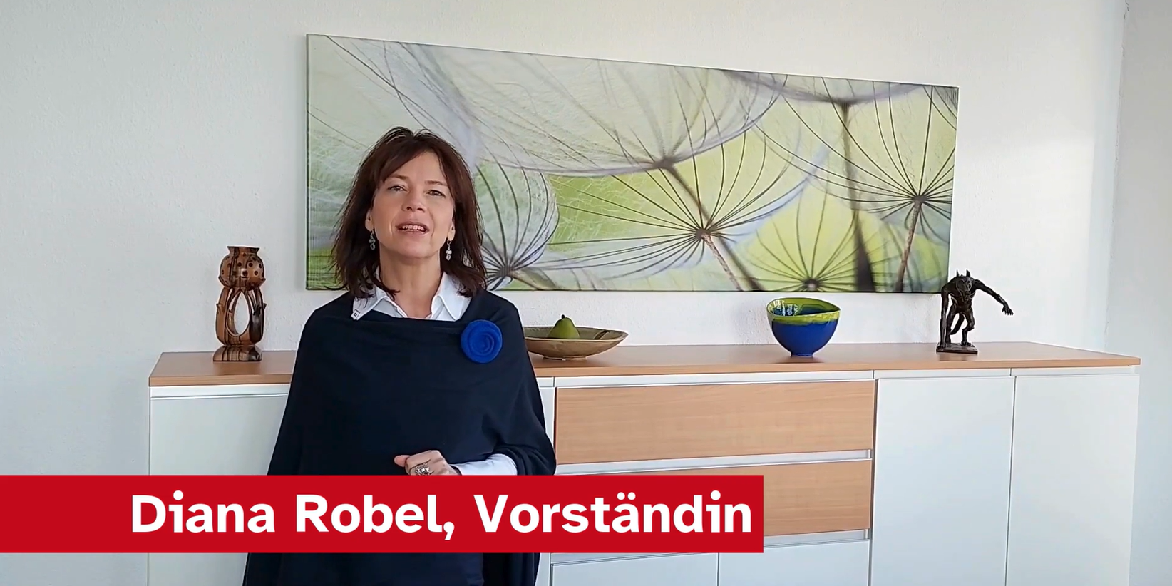 Eine Frau steht vor einem modernen Sideboard, auf dem Dekorationsobjekte und eine Schüssel stehen. Im Hintergrund ist ein großes, farbenfrohes Bild von Pflanzen zu sehen. Sie trägt einen eleganten schwarzen Umhang und lächelt. Am unteren Rand steht der Text: „Diana Robel, Vorstandin“.