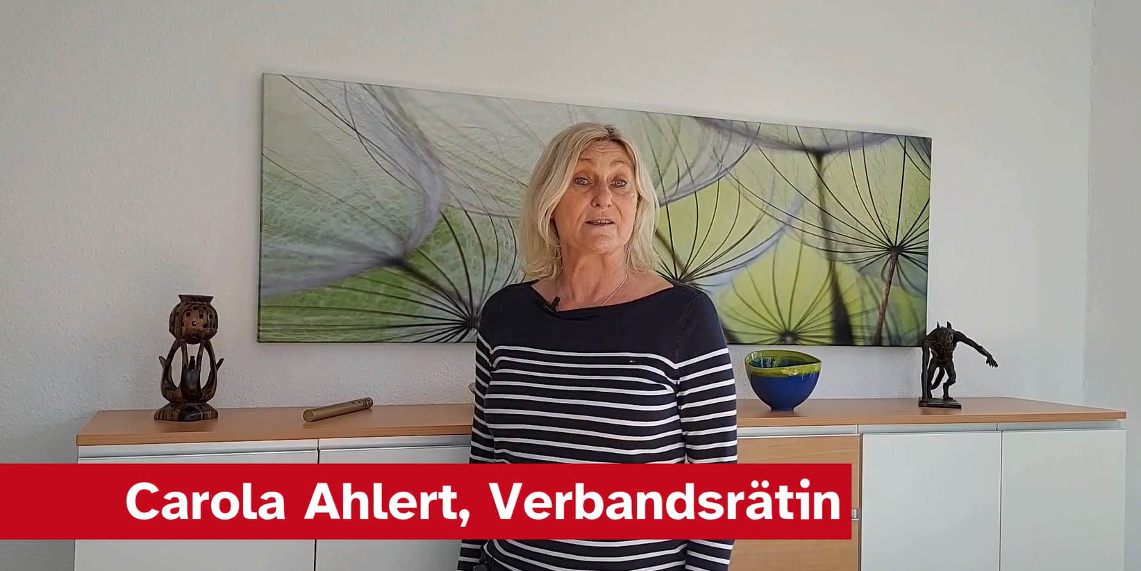 Eine Frau mit langen, blonden Haaren steht vor einer hellen Wand. Sie trägt ein gestreiftes Oberteil und spricht in die Kamera, während im Hintergrund ein modernes Gemälde sowie einige Dekorationsobjekte auf einem Sideboard sichtbar sind.